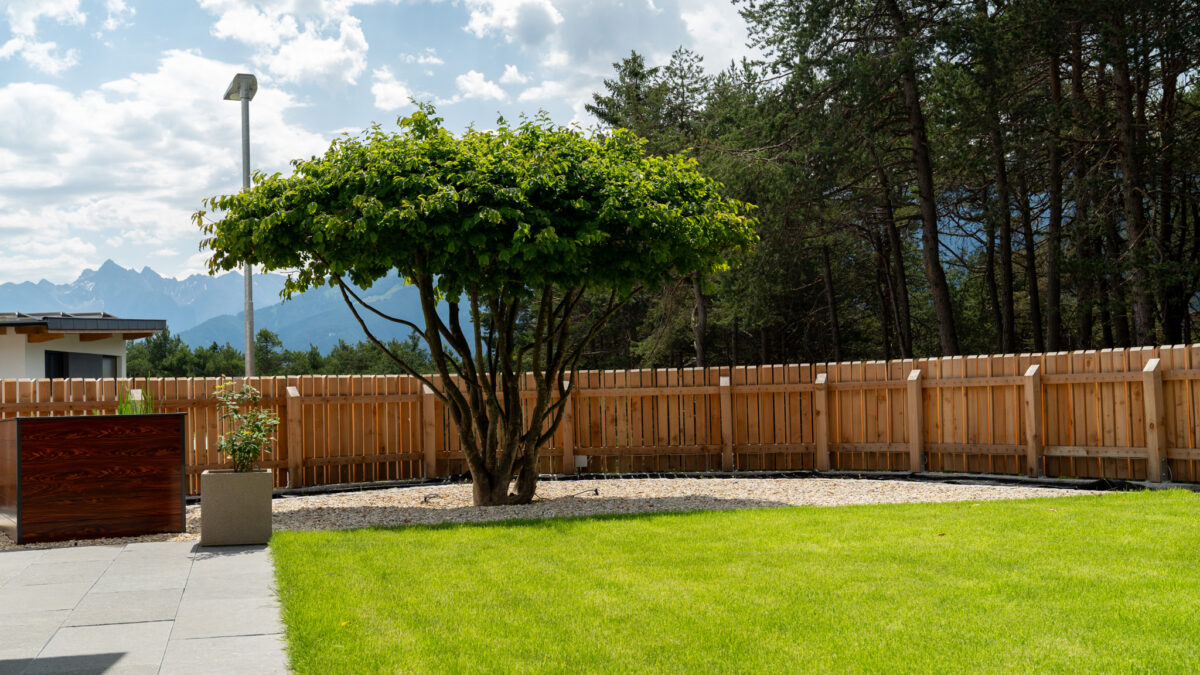 grasberger fließerau Ein üppig grüner Garten in Tirol von Grasberger Fliesserau, mit getrimmtem Rasen, einem zentralen Baum, der von Kies umgeben ist, einem Holzzaun, Topfpflanzen und Bäumen im Hintergrund unter einem teilweise bewölkten Himmel - entworfen vom Experten Gärtner.