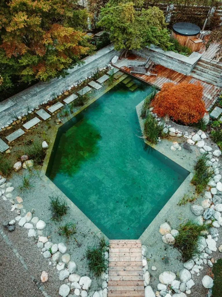 grasberger fließerau Ein von Grasberger Gärtner gestalteter Naturpool mit klarem, grünem Wasser ist von Steinen, Pflanzen und einer Holzterrasse umgeben. Eine Treppe führt in den Pool in der Nähe eines kleinen Stegs und Liegestühlen, während Bäume und Sträucher ein ruhiges Tiroler Gartenambiente schaffen.