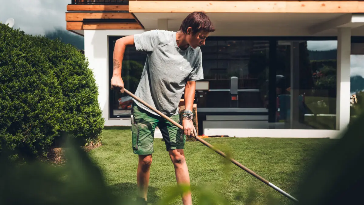 grasberger fließerau Eine Person in grauem T-Shirt und grünen Shorts ist mit der Gartenpflege beschäftigt und arbeitet im Rasen hinter einem von Büschen und Grün umgebenen Garten, mit einem modernen Haus und großen Fenstern im Hintergrund.