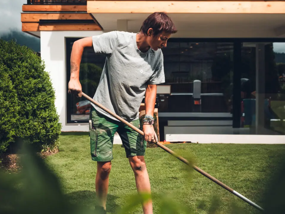 grasberger fließerau Eine Person in grauem T-Shirt und grünen Shorts ist mit der Gartenpflege beschäftigt und arbeitet im Rasen hinter einem von Büschen und Grün umgebenen Garten, mit einem modernen Haus und großen Fenstern im Hintergrund.