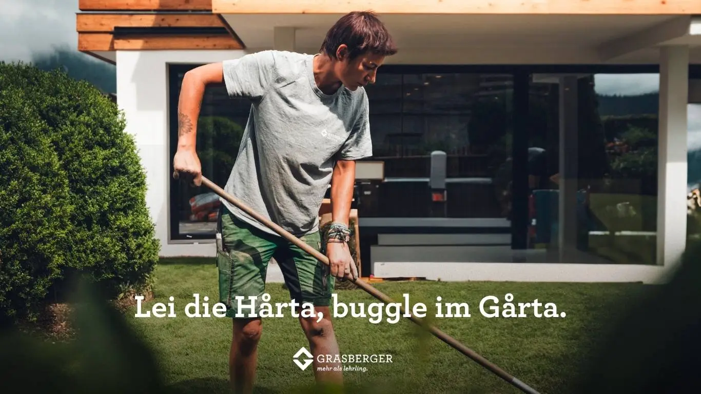 grasberger fließerau Eine Person in grauem T-Shirt und Shorts harkt das Gras in einem Garten, der in der Nähe eines modernen Hauses steht. Der Text lautet: "Lei die Härta, buggle im Gårta". Unten ist das Grasberger-Logo zu sehen - Experten für Gartenbau aus Landeck, Tirol.