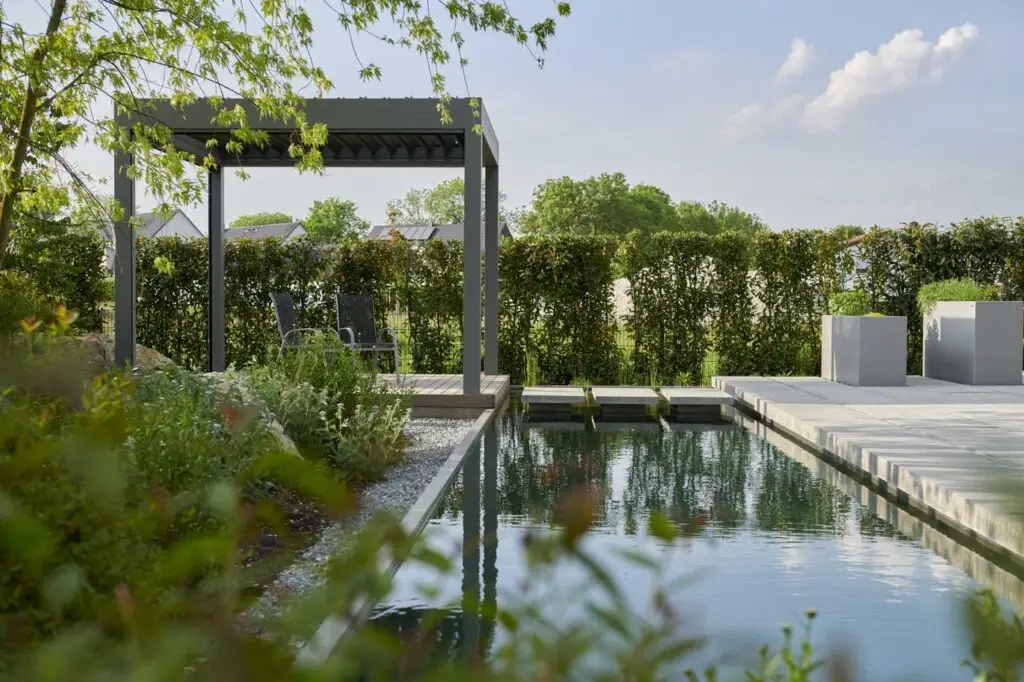 grasberger fließerau Moderner Garten mit einem rechteckigen Teich, Trittsteinen, hohen Hecken und einem schattigen Sitzplatz unter einer Pergola - ein einladender Rückzugsort inmitten üppigen Grüns und inspiriert von Gartentrends 2026 an einem sonnigen Tag.