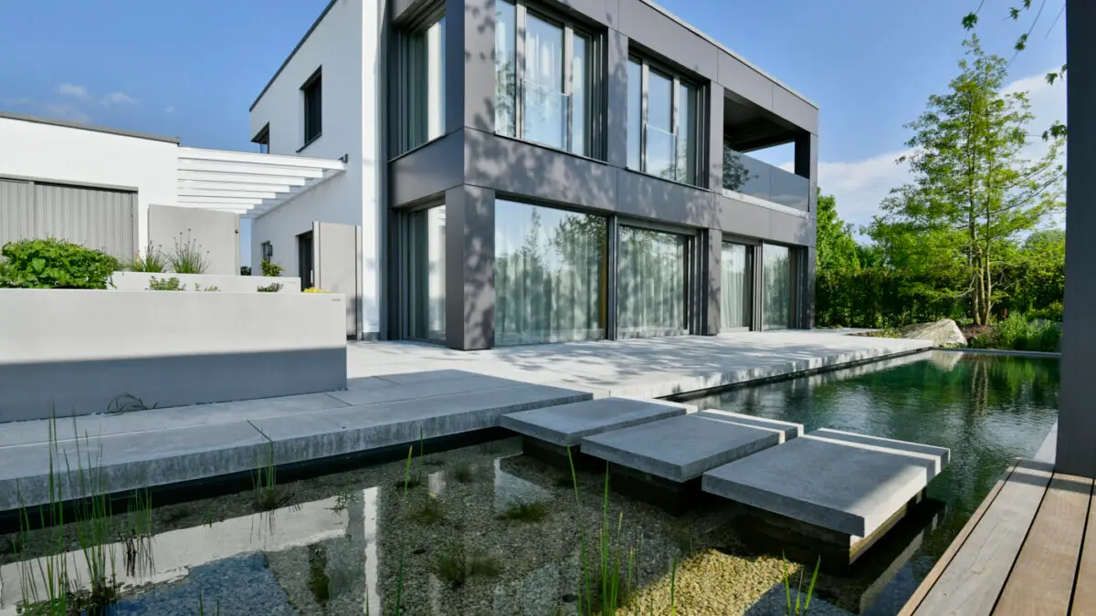 grasberger fließerau Ein modernes zweistöckiges Haus mit großen Fenstern, grau-weißem Äußeren und minimalistischem Design. Die Szene, die Gartentrends 2026 präsentiert, zeigt einen Teich mit Trittsteinen, üppige Pflanzen im Vordergrund und einen klaren Himmel darüber.