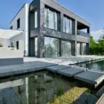 grasberger fließerau Ein modernes zweistöckiges Haus mit großen Fenstern, grau-weißem Äußeren und minimalistischem Design. Die Szene, die Gartentrends 2026 präsentiert, zeigt einen Teich mit Trittsteinen, üppige Pflanzen im Vordergrund und einen klaren Himmel darüber.