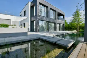 grasberger fließerau Ein modernes zweistöckiges Haus mit großen Fenstern, grau-weißem Äußeren und minimalistischem Design. Die Szene, die Gartentrends 2026 präsentiert, zeigt einen Teich mit Trittsteinen, üppige Pflanzen im Vordergrund und einen klaren Himmel darüber.