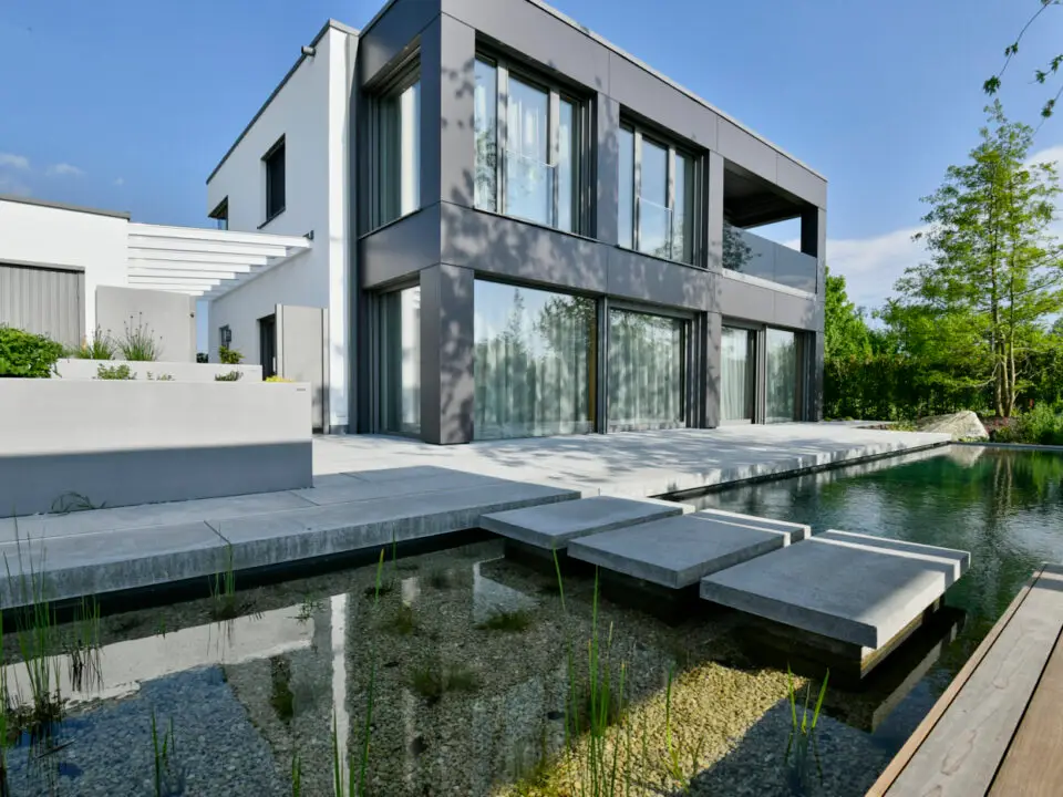 grasberger fließerau Ein modernes zweistöckiges Haus mit großen Fenstern, grau-weißem Äußeren und minimalistischem Design. Die Szene, die Gartentrends 2026 präsentiert, zeigt einen Teich mit Trittsteinen, üppige Pflanzen im Vordergrund und einen klaren Himmel darüber.