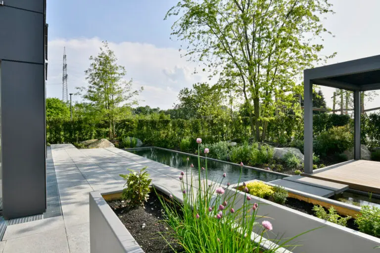 grasberger fließerau Ein moderner Garten im Hinterhof, inspiriert von Gartentrends 2026, bietet Hochbeete, einen Teich, üppiges Grün und eine Pergola an einem sonnigen Tag. Betonwege umrahmen den Raum, während hohe Bäume einen Teil des Schattens für die Entspannung im Freien bieten.