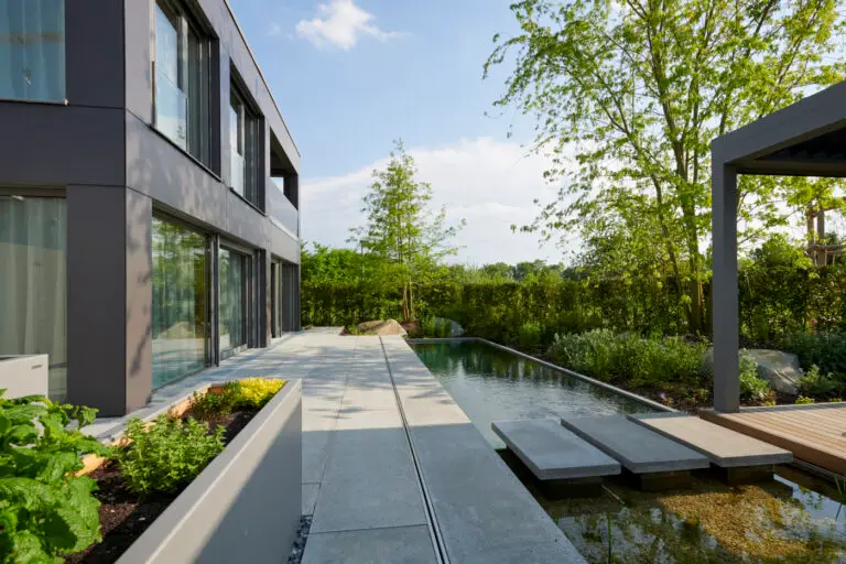 grasberger fließerau Modernes Haus mit großen Fenstern und einem Innenhof neben einem schmalen Gartenteich. Trittsteine überqueren das Wasser, umgeben von üppigen grünen Pflanzen und Bäumen unter einem klaren blauen Himmel - ein perfektes Schaufenster für Gartentrends 2026.