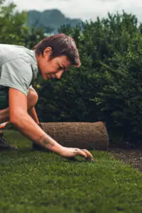 grasberger fließerau Eine Person kniet auf dem Boden und legt eine Rolle Rollrasen auf einem Rasen mit grünen Sträuchern im Hintergrund aus. Mit kurzen Haaren und einem Tattoo auf dem Unterarm ist sie auf ihre Arbeit konzentriert - vielleicht setzt sie neue Gartentrends 2026 im Landschaftsbau.